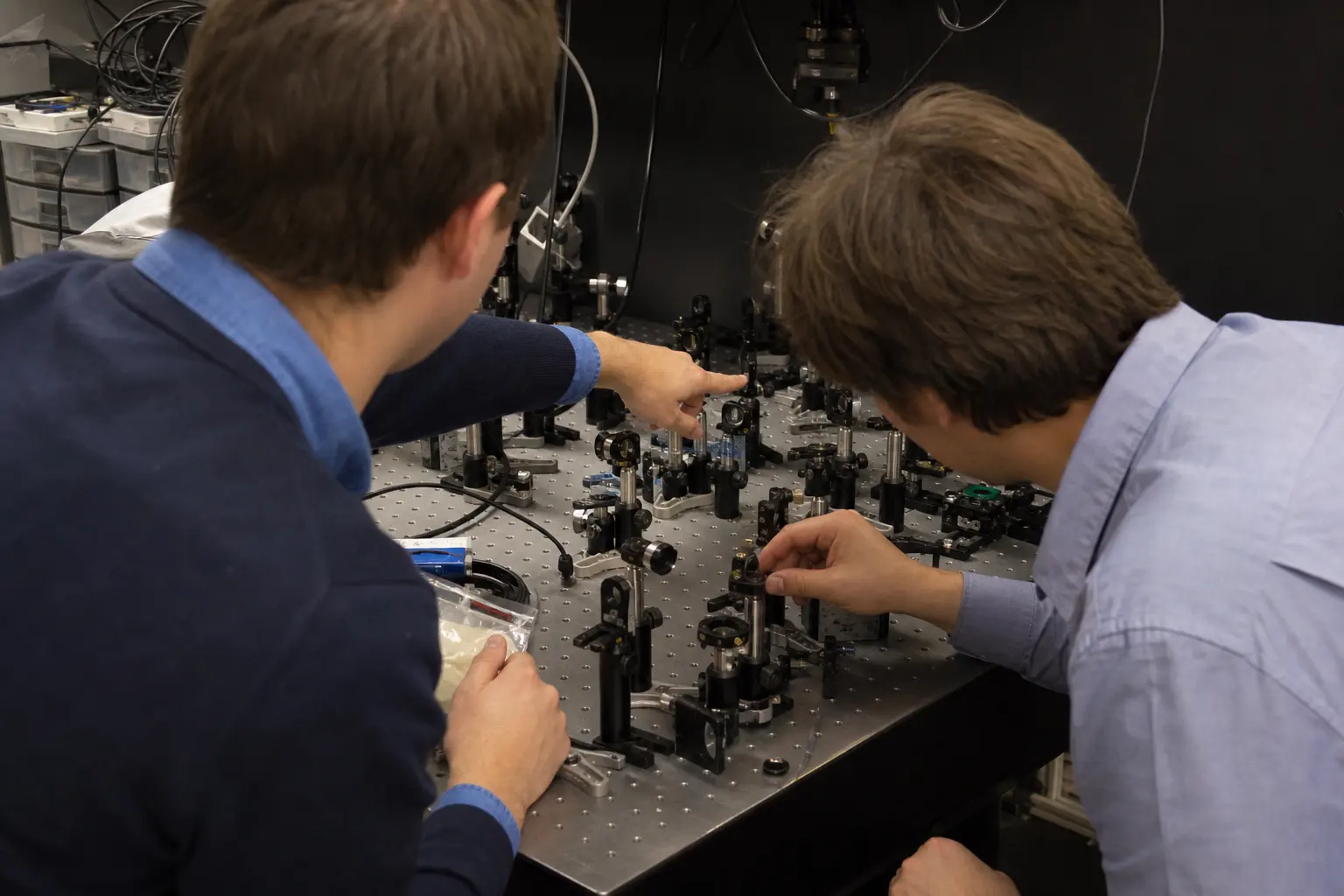 Two engineers at an optics bench crowded with mounts, mirrors and cabling, one pointing at the setup while the other adjusts a mount